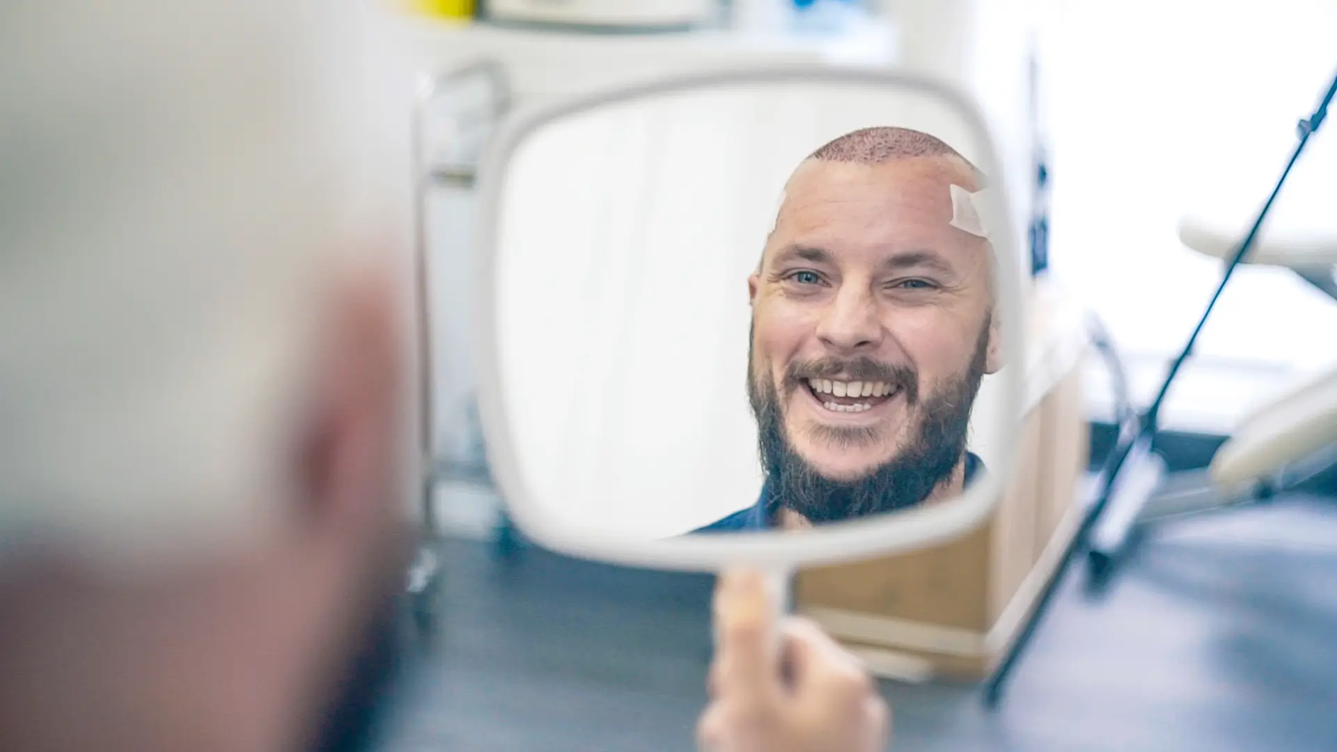 A smiling bald man with a beard observing his hair regrowth in a handheld mirror in a bright, modern room, possibly a barbershop or salon.