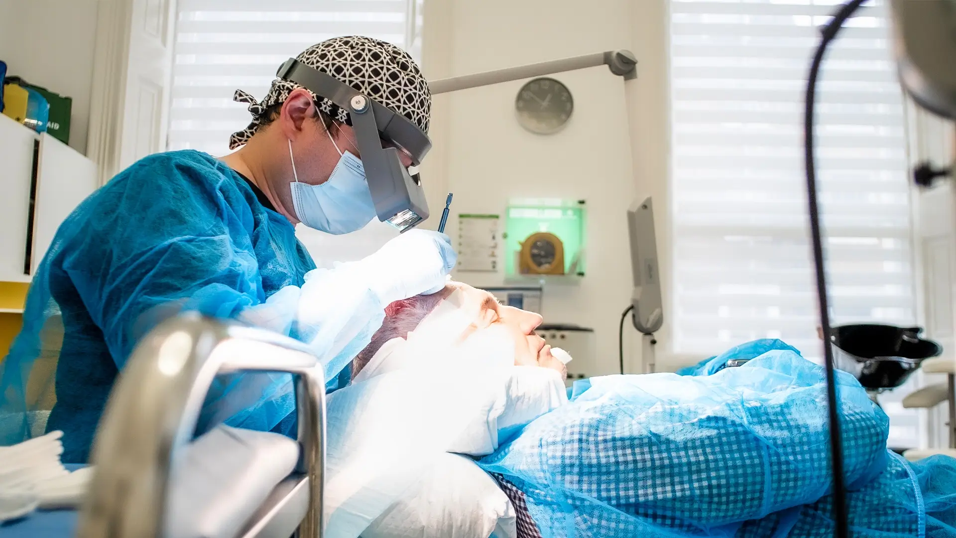 A dentist in blue scrubs and protective gear is examining a patient's teeth in a well-equipped dental office. The patient, dealing with female hair loss, is lying back in a dental chair, covered