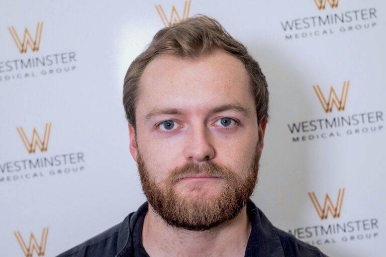 A man with a beard and short hair, possibly looking into hair transplant services, stands in front of a backdrop with the Westminster Medical Group logo repeated in a pattern.