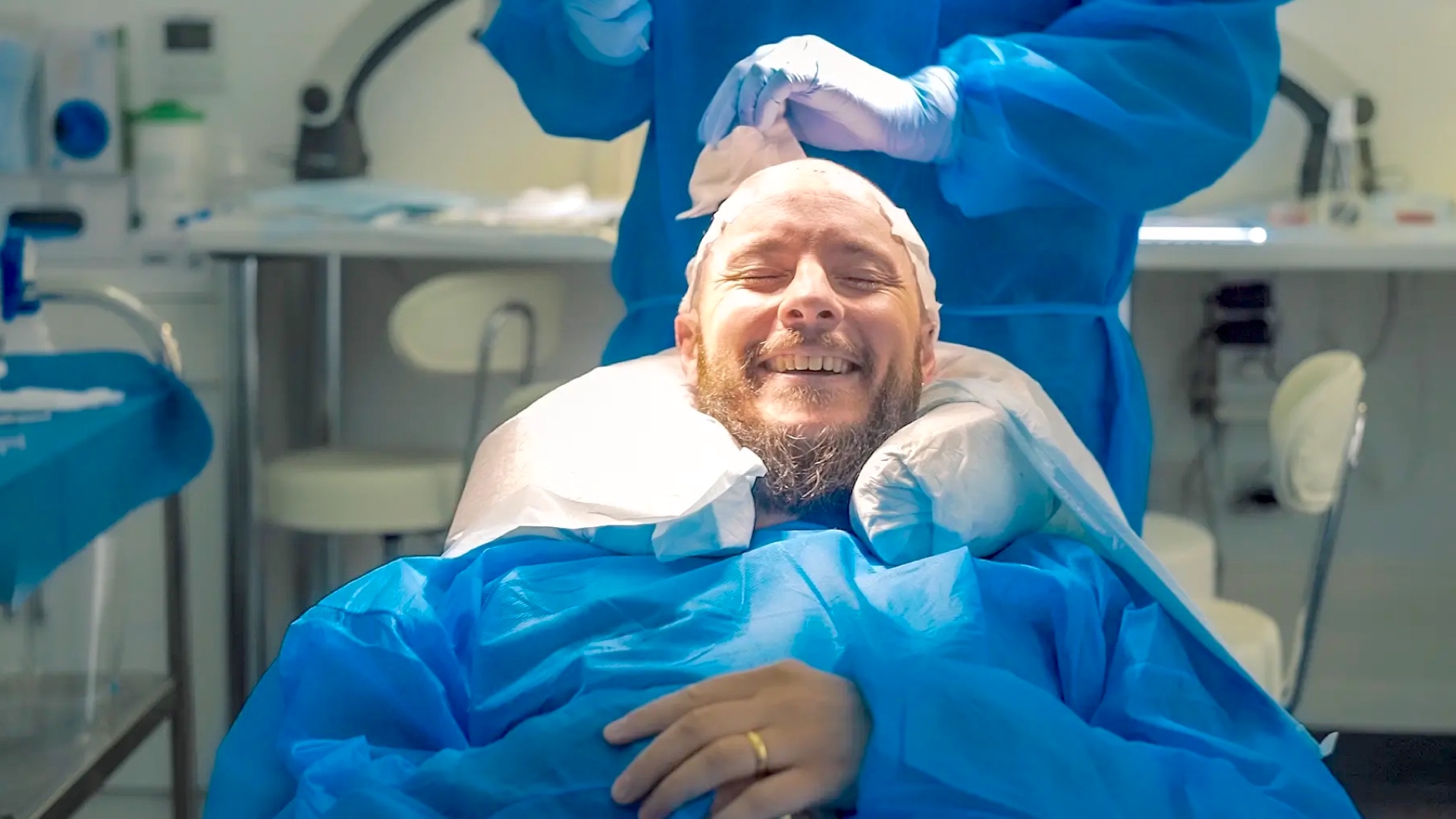 A smiling man with a beard, wearing a surgical cap and covered with a blue surgical drape, lies in an operating room with medical staff attending to him for a hair implant procedure.