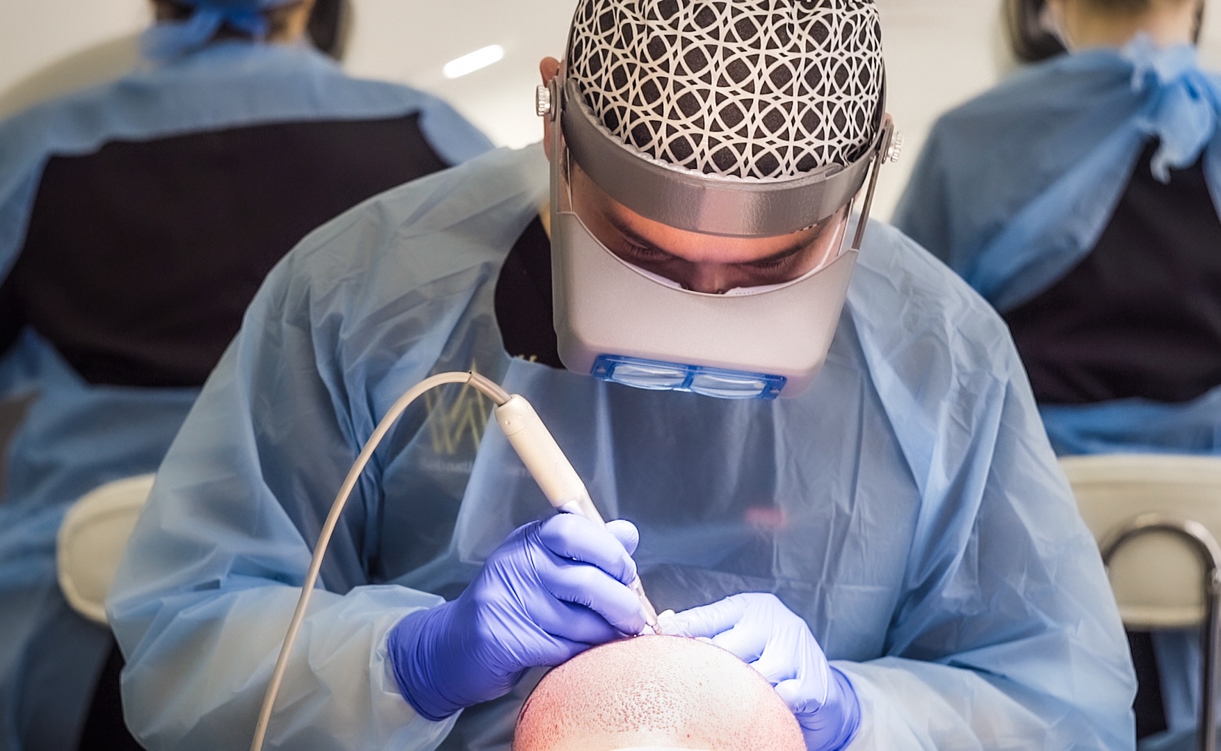 A medical professional specializing in hair surgery, wearing a surgical cap, face shield, and gloves, performs a procedure for male pattern baldness, concentrating intensely on the patient's head in a clinical setting.