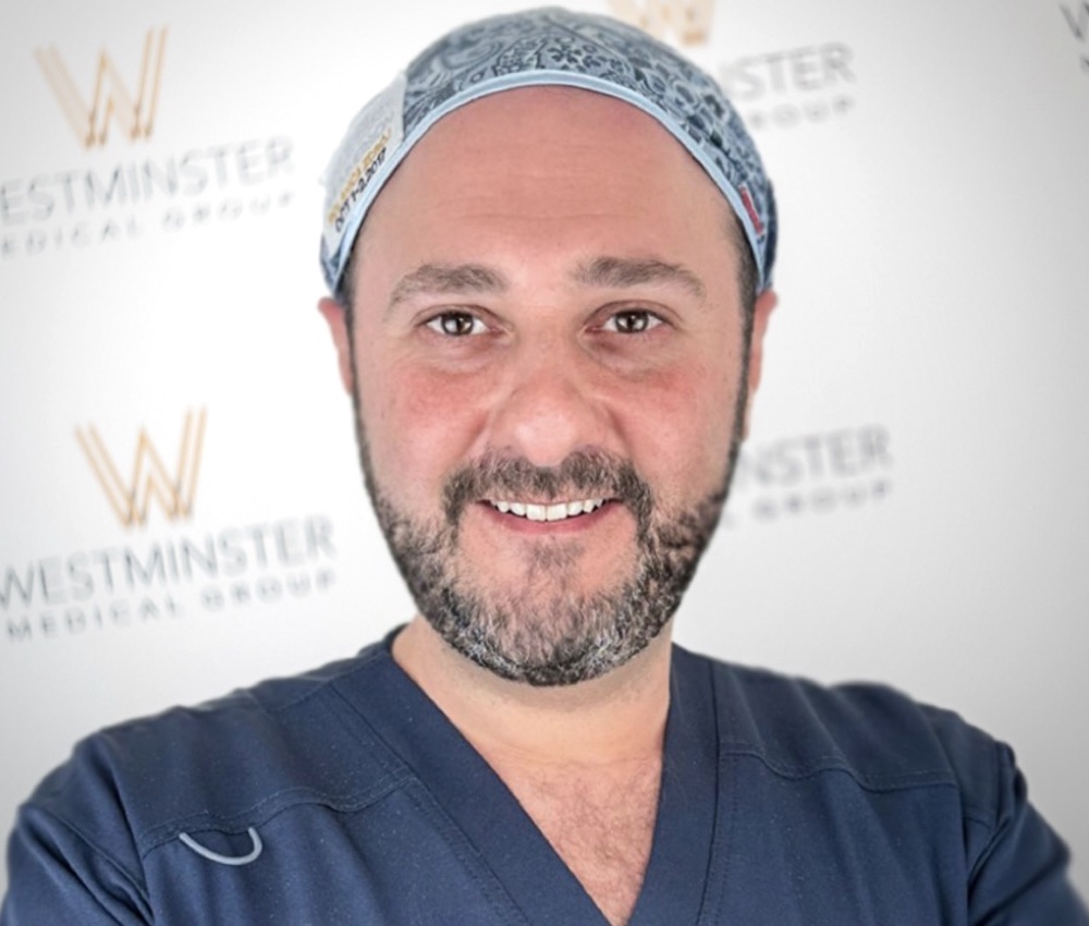 A smiling man wearing medical scrubs and a patterned scrub cap stands in front of a banner with the "Westminster Medical Group" logo, specializing in hair surgery.