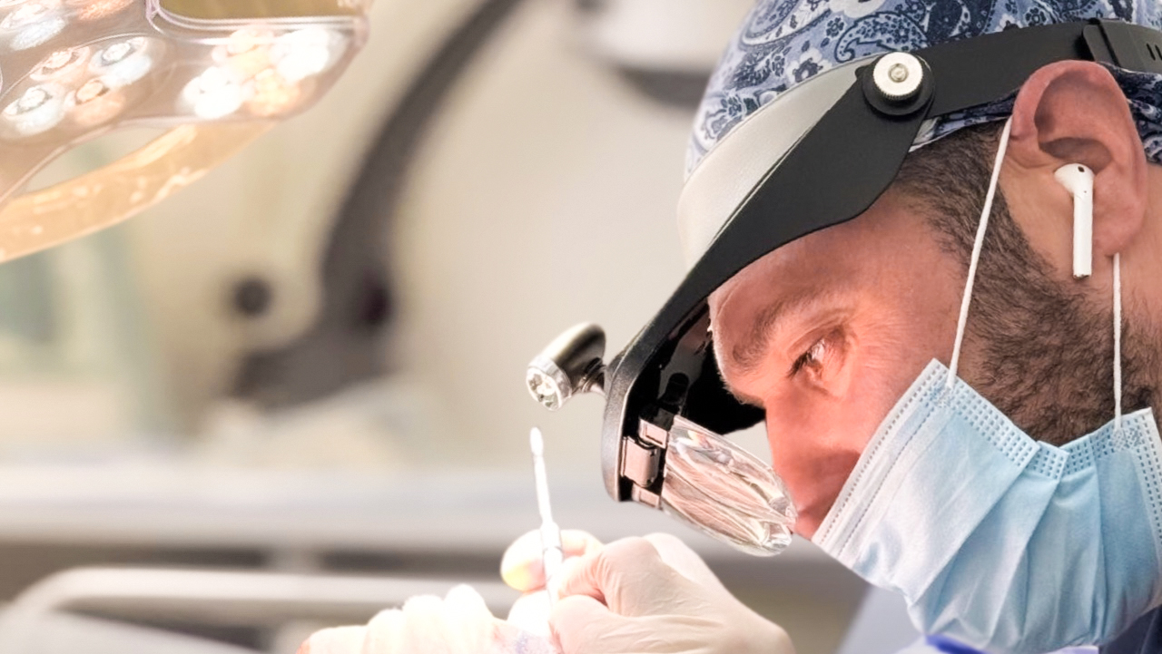 A focused surgeon wearing surgical loupes and a mask performs a precise hair replacement procedure under bright operating lights, with medical tools in hand.