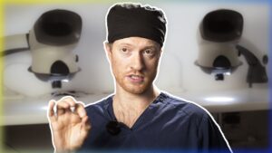 A male surgeon in blue scrubs and a black head covering talks to the camera, with blurred images of surgical lights in the background, specializing in hair implants.