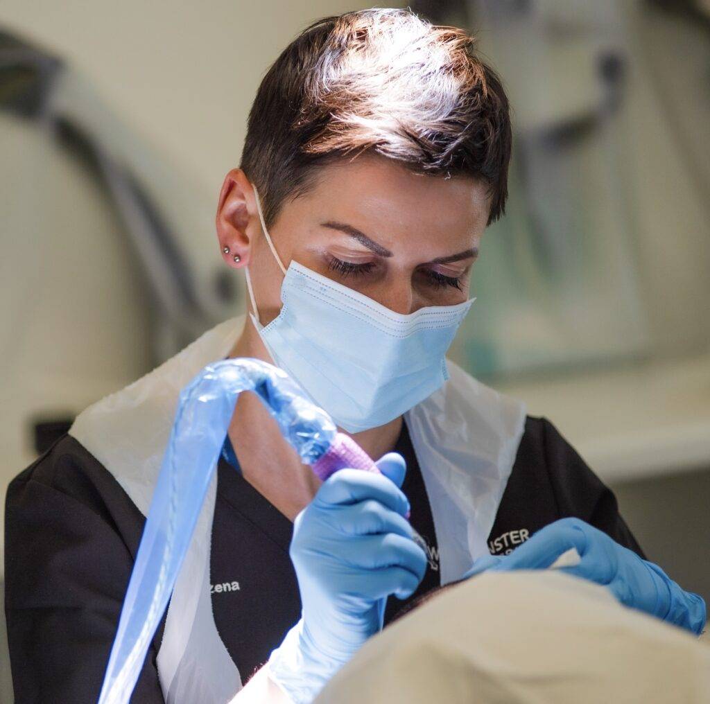 A focused female dentist with short hair, wearing a mask, gloves, and a protective apron, is performing a dental procedure on a patient experiencing female hair loss.